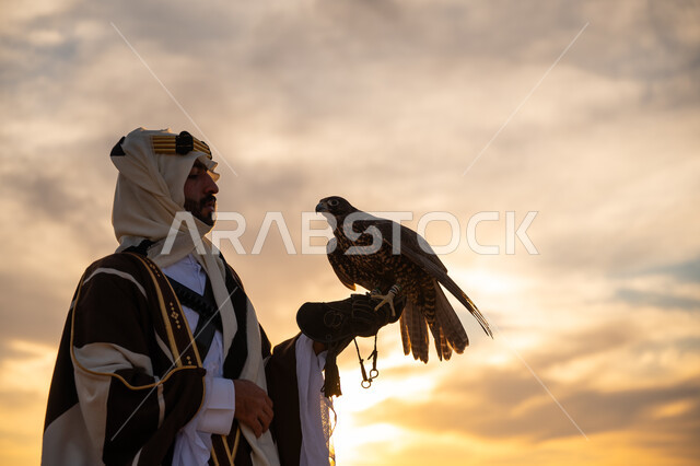 ذكرى تأسيس الدوله السعوديه الاولى 1727 م, اهمية الصقور في التاريخ السعودي, رمز القوة والتحدي وحدة البصر, توارث شغف تربية الجوارح, منظر السماء عند شروق الشمس, يوم بدينا 22 فبراير، صورة مقربة من الجانب لرجل عربي خليجي سعودي يرتدي البشت والعقال يحمل طائر الصقر