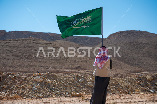 يوم العلم 11 مارس, ذكرى تأسيس الدولة السعودية الأولى في 1727م، صورة من الخلف لرجل عربي خليجي سعودي يرتدي الثوب التقليدي والشماغ يقف في الصحراء يحمل علم المملكة العربية السعودية، راية الإسلام والتوحيد، يوم بدينا 22 فبراير، ارتداء الزي الشعبي في المناسبة الوطنية