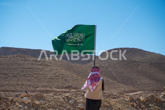 راية الإسلام والتوحيد، يوم العلم 11 مارس, ذكرى تأسيس الدولة السعودية الأولى في 1727م، صورة من الخلف لرجل عربي خليجي سعودي يرتدي الثوب التقليدي والشماغ يقف في الصحراء يحمل علم المملكة العربية السعودية، يوم بدينا 22 فبراير، ارتداء الزي الشعبي في المناسبة الوطنية