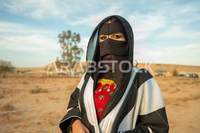 ارتداء زي المنطقة الوسطى في المناسبة الوطنية، يوم بدينا 22 فبراير، الاهتمام والعناية بالمظهر الخارجي، ذكرى تأسيس الدولة السعودية الأولى 1727 م، صورة مقربة لإمرأة خليجية عربية سعودية ترتدي البشت النسائي السعودي والبرقع تقف على الرمال الذهبية الناعمة في الصحراء