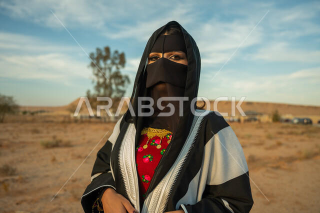 ارتداء زي المنطقة الوسطى في المناسبة الوطنية، يوم بدينا 22 فبراير، الاهتمام والعناية بالمظهر الخارجي، ذكرى تأسيس الدولة السعودية الأولى 1727 م، صورة مقربة لإمرأة خليجية عربية سعودية ترتدي البشت النسائي السعودي والبرقع تقف على الرمال الذهبية الناعمة في الصحراء
