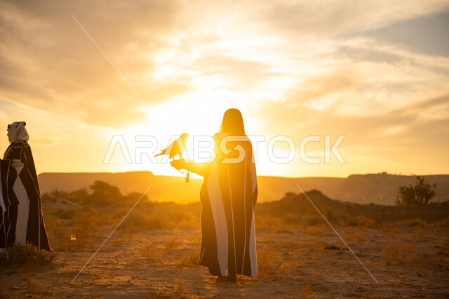 The status of falcons in Saudi history, the anniversary of the founding of the first Saudi state 1727 AD, the day we started February 22, sunrise in the desert nature, inheriting the passion for raising birds of prey, a symbol of strength, challenge and sharp eyesight, a Saudi Gulf Arab man and woman wearing traditional dress and carrying a falcon