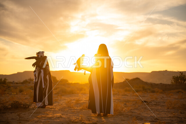 ذكرى تأسيس الدوله السعوديه الاولى 1727 م, يوم بدينا 22 فبراير، غروب الشمس في الصحراء, توارث شغف تربية الجوارح, رمز القوة والتحدي وحدة البصر, اهمية الصقور في التاريخ السعودي, رجل وامرأة عربيان خليجيان سعوديان يرتديان الزي الشعبي ويحملان طائر الصقر