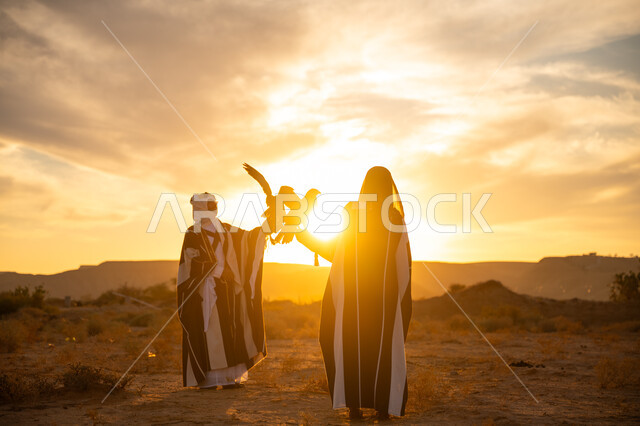 رمز القوة والتحدي وحدة البصر, اهمية الصقور في التاريخ السعودي, ذكرى تأسيس الدوله السعوديه الاولى 1727 م, يوم بدينا 22 فبراير، توارث شغف تربية الجوارح, رجل وامرأة عربيان خليجيان سعوديان يرتديان الزي الشعبي ويحملان طائر الصقر, غروب الشمس في الصحراء