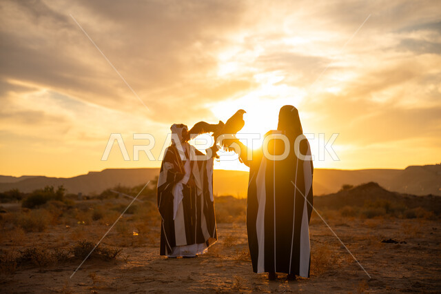 Anniversary of the founding of the first Saudi state 1727 AD, the day we started February 22, the importance of falcons in Saudi history, a symbol of strength, challenge and sharp eyesight, inheritance of the passion for raising birds of prey, a Saudi Gulf Arab man and woman wearing traditional dress and carrying a falcon, sunrise in the desert nature