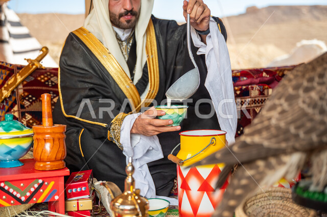 Wearing traditional dress on the founding day of the first Saudi state, February 22, authentic Arab customs to celebrate a national holiday, a Saudi Gulf Arab man wearing a bisht, ghutra and a wide golden agal pouring milk into a colorful ceramic pot, the day of our beginning 1727 AD, a popular handmade product, raising a raptor