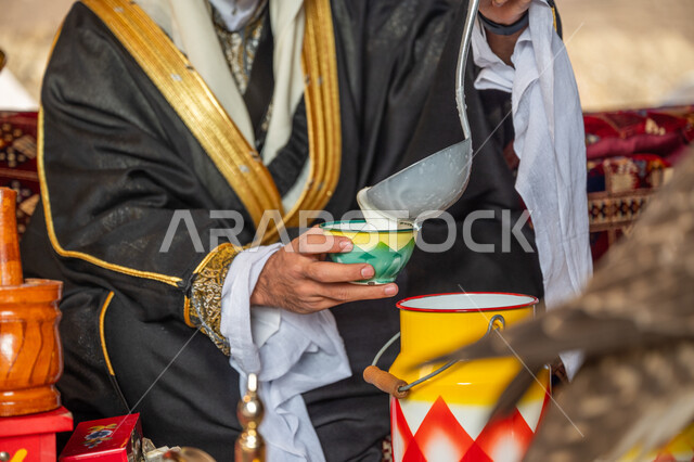 يوم بدينا 1727 م، العادات العربية الأصيلة، المنتج اليدوي الشعبي، ارتداء الزي الشعبي في يوم تأسيس الدولة السعودية الأولى 22 فبراير, الاحتفال بعيد ومناسبة وطنية, صورة مقربة لرجل عربي خليجي سعودي يرتدي البشت والغترة يقوم بصب اللبن في إناء خزفي ملون
