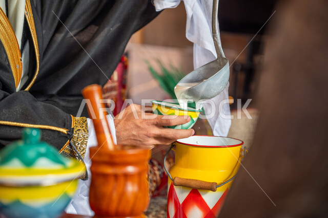 المنتج اليدوي الشعبي، ارتداء الزي الشعبي في يوم تأسيس الدولة السعودية الأولى 22 فبراير, العادات العربية الأصيلة، الاحتفال بعيد ومناسبة وطنية, صورة مقربة لرجل عربي خليجي سعودي يرتدي البشت والغترة يقوم بصب اللبن في إناء خزفي ملون, يوم بدينا 1727 م