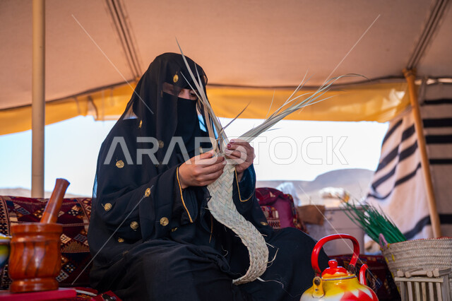 الفخر بالتراث السعودي العريق، يوم بدينا 22 فبراير، امرأة عربية خليجية سعودية ترتدي العباءة المطرزة والبرقع تجلس في الخيمة وتقوم بصناعة الحصير والسلل من الخوص الجاف، عرض المنتجات المحليه الوطنيه، حرفة يدوية شعبية، ذكرى تأسيس الدولة السعودية الأولى 1727 م