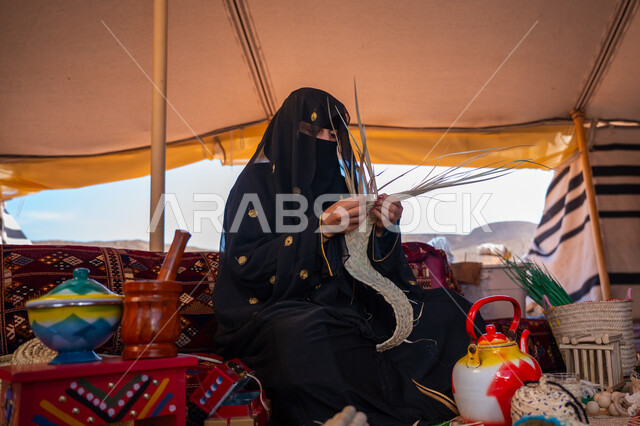 حرفة يدوية شعبية، الفخر بالتراث السعودي العريق، يوم بدينا 22 فبراير، امرأة عربية خليجية سعودية ترتدي العباءة المطرزة والبرقع تجلس في الخيمة وتقوم بصناعة الحصير والسلل من الخوص الجاف، عرض المنتجات المحليه الوطنيه، ذكرى تأسيس الدولة السعودية الأولى 1727 م