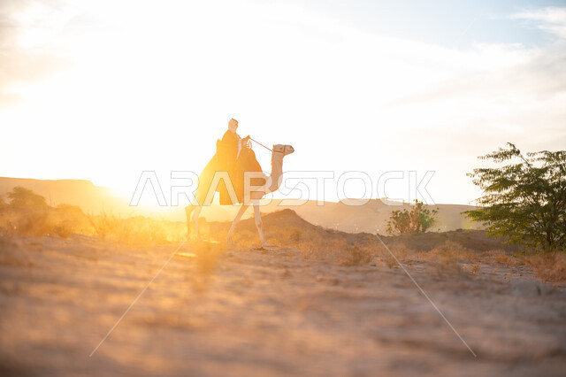 ارتداء الزي الشعبي في المناسبة الوطنية, صورة من الجانب لرجل عربي خليجي سعودي يرتدي البشت التقليدي والغترة يمتطي الجمل وسط الصحراء في وقت غروب الشمس, ذكرى تأسيس الدولة السعودية الأولى 1727م, الاعتزاز والفخر بالانتماء للوطن, يوم بدينا 22 فبراير