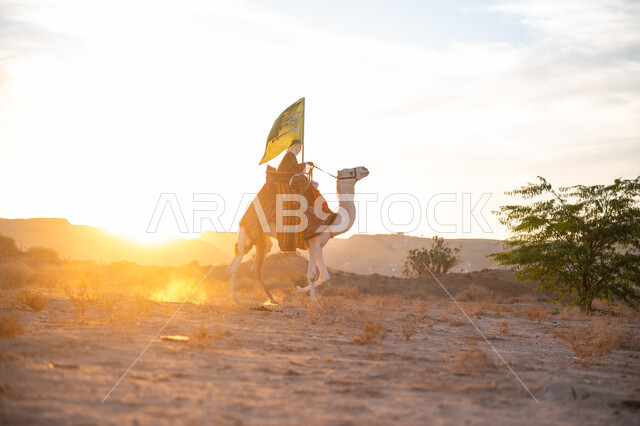 ذكرى تأسيس الدولة السعودية الأولى  1727م, ارتباط الإبل بالأصالة والعراقة, ارتداء الزي الشعبي في المناسبة الوطنية، يوم بدينا 22 فبراير, صورة من الجانب لرجل عربي خليجي سعودي يرتدي البشت التقليدي والغترة يمتطي الجمل وسط الصحراء ويحمل علم المملكة وقت غروب الشمس