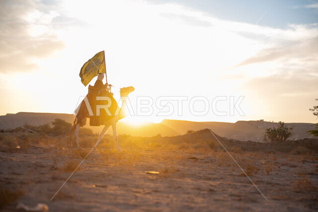 ذكرى تأسيس الدولة السعودية الأولى  1727م, ارتباط الإبل بالأصالة والعراقة, ارتداء الزي الشعبي في المناسبة الوطنية، يوم بدينا 22 فبراير, صورة من الجانب لرجل عربي خليجي سعودي يرتدي البشت التقليدي والغترة يمتطي الجمل وسط الصحراء ويحمل علم المملكة وقت غروب الشمس