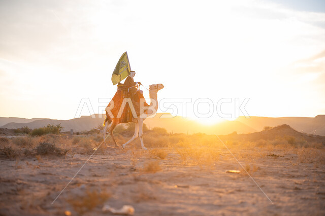 The association of camels with authenticity and antiquity, wearing traditional costume on the national occasion, the day of our beginning, February 22, a side view of a Saudi Arabian Gulf man wearing the traditional bisht and ghutra riding a camel in the middle of the desert and carrying the flag of the Kingdom at sunset, the anniversary of the founding of the first Saudi state 1727 AD
