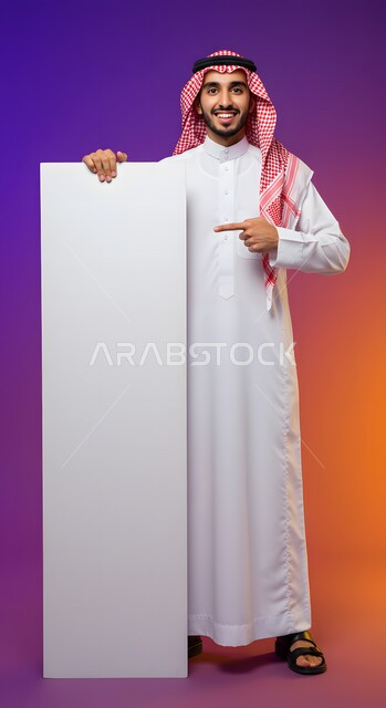 استخدام لوحة توضيحية ووسيلة تعليمية، إعلان وعرض تسويقي، موك اب مربع أبيض، بورتريه لشاب عربي خليجي سعودي مبتسم يرتدي الثوب التقليدي والشماغ يشير بيده إلى لوحه بيضاء مربعه فارغة ينظر إلى الكاميرا بإيماءات السرور، صورة الجسم بالطول الكامل، خلفية ملونة