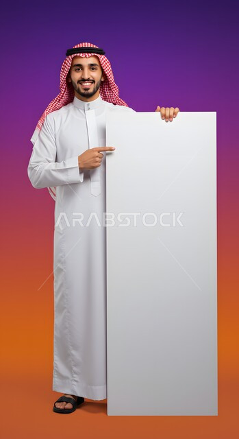 موك اب مربع أبيض، إعلان وعرض تسويقي، استخدام لوحة توضيحية ووسيلة تعليمية، بورتريه لشاب عربي خليجي سعودي مبتسم يرتدي الثوب التقليدي والشماغ يشير بإصبع السبابة إلى لوحه بيضاء مربعه فارغة ينظر إلى الكاميرا بإيماءات السرور، صورة الجسم بالطول الكامل، خلفية ملونة