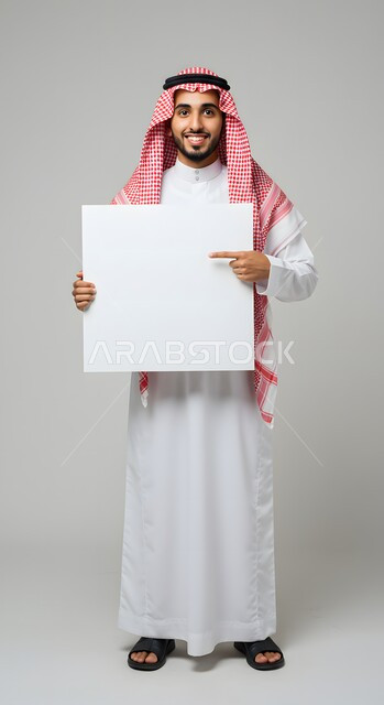 موك اب مربع أبيض، استخدام لوحة توضيحية ووسيلة تعليمية، عرض وإعلان تسويقي، بورتريه لشاب عربي خليجي سعودي مبتسم يرتدي الثوب التقليدي والشماغ يشير بإصبع السبابة الى لوحه بيضاء مربعه فارغة ينظر إلى الكاميرا بإيماءات السرور، صورة الجسم بالطول الكامل، خلفية ملونة