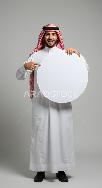 استخدام لوحة توضيحية ووسيلة تعليمية، إعلان وعرض تسويقي، موك اب دائري أبيض، بورتريه لشاب عربي خليجي سعودي مبتسم يرتدي الثوب التقليدي والشماغ يشير بإصبع السبابة الى لوحه بيضاء مدورة فارغة ينظر إلى الكاميرا بإيماءات السرور، صورة الجسم بالطول الكامل، خلفية ملونة
