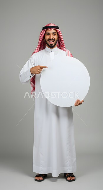 موك اب دائري أبيض، استخدام لوحة توضيحية ووسيلة تعليمية، إعلان وعرض تسويقي، بورتريه لشاب عربي خليجي سعودي مبتسم يرتدي الثوب التقليدي والشماغ يشير بإصبع السبابة الى لوحه بيضاء مدورة فارغة ينظر إلى الكاميرا بإيماءات السرور، صورة الجسم بالطول الكامل، خلفية ملونة