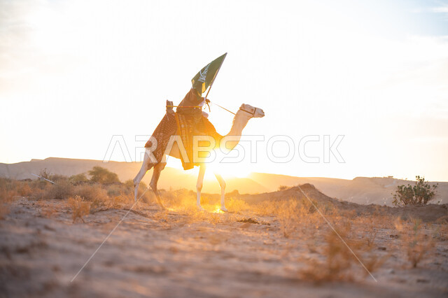 يوم بدينا 22 فبراير, ذكرى تأسيس الدولة السعودية الأولى  1727م, ارتباط الإبل بالأصالة والعراقة, ارتداء الزي الشعبي في المناسبة الوطنية, رجل عربي خليجي سعودي يرتدي البشت والغترة يمتطي الجمل في الصحراء ويحمل علم المملكة وقت غروب الشمس, الفخر بالإنتماء الى الوطن