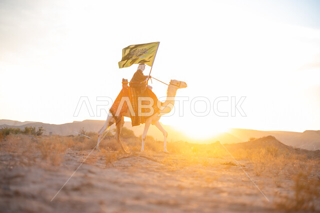 يوم بدينا 22 فبراير, ذكرى تأسيس الدولة السعودية الأولى  1727م, ارتباط الإبل بالأصالة والعراقة, ارتداء الزي الشعبي في المناسبة الوطنية, رجل عربي خليجي سعودي يرتدي البشت والغترة يمتطي الجمل في الصحراء ويحمل علم المملكة وقت غروب الشمس, الفخر بالإنتماء الى الوطن