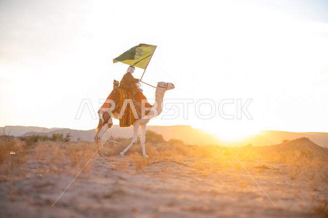The association of camels with authenticity and antiquity, wearing traditional dress on a national occasion, a Saudi Gulf Arab man wearing a bisht and a ghutra riding a camel in the desert and carrying the flag of the Kingdom at sunset, pride in belonging to the homeland, the day we started, February 22, the anniversary of the founding of the first Saudi state 1727 AD