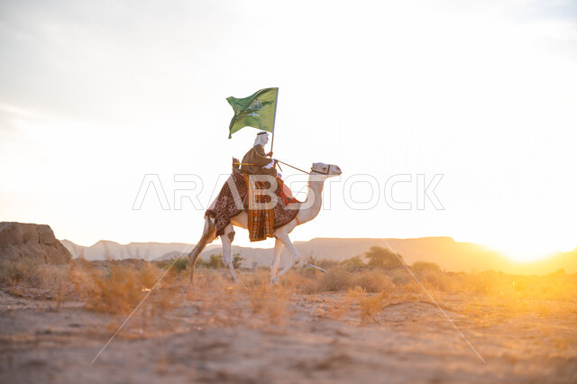 يوم بدينا 22 فبراير, ذكرى تأسيس الدولة السعودية الأولى  1727م, ارتباط الإبل بالأصالة والعراقة, ارتداء الزي الشعبي في المناسبة الوطنية, رجل عربي خليجي سعودي يرتدي البشت والغترة يمتطي الجمل في الصحراء ويحمل علم المملكة وقت غروب الشمس, الفخر بالإنتماء الى الوطن