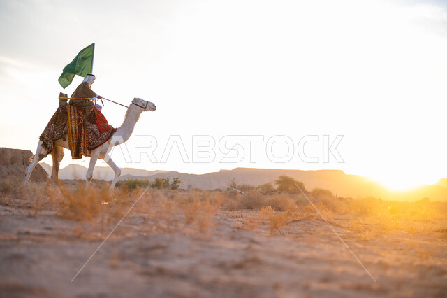 ذكرى تأسيس الدولة السعودية الأولى  1727م, ارتباط الإبل بالأصالة والعراقة, ارتداء الزي الشعبي في المناسبة الوطنية, رجل عربي خليجي سعودي يرتدي البشت والغترة يمتطي الجمل في الصحراء ويحمل علم المملكة وقت غروب الشمس, الفخر بالإنتماء الى الوطن, يوم بدينا 22 فبراير