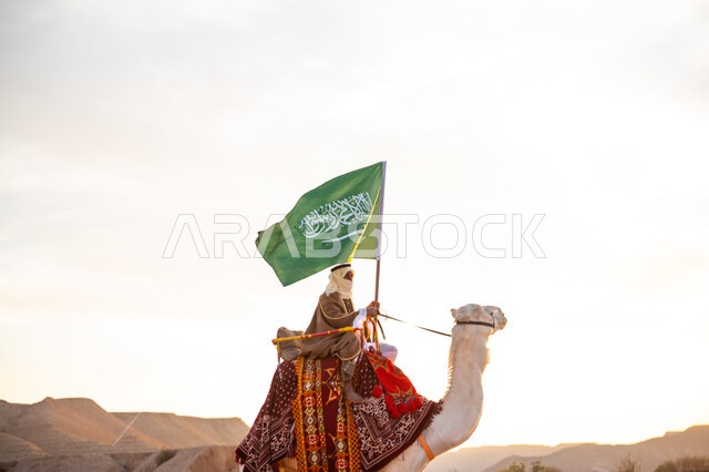 الاحتفال بعيد ومناسبة وطنية, ارتداء الزي الشعبي بيوم تأسيس الدولة السعودية الأولى 1727 م, يوم بدينا 22 فبراير, الفخر والاعتزاز بالإنتماء الى الوطن، ارتباط الإبل بالتراث السعودي, رجل عربي خليجي سعودي يرتدي البشت والغترة يمتطي الجمل في الصحراء ويحمل علم المملكة