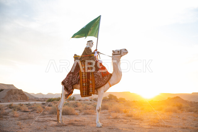 يوم بدينا 22 فبراير, الفخر والإنتماء للوطن، ارتباط الإبل بالتراث السعودي, رجل عربي خليجي سعودي يرتدي البشت والغترة يمتطي الجمل في الصحراء ويحمل علم المملكة بوقت الغروب, ارتداء الزي الشعبي بيوم تأسيس الدولة السعودية الأولى 1727 م, الاحتفال بعيد ومناسبة وطنية