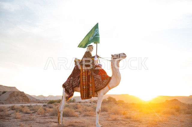 Wearing the traditional costume on the founding day of the first Saudi state 1727 AD, the day we started on February 22, pride and belonging to the homeland, the connection of camels to Saudi heritage, a Saudi Gulf Arab man wearing a bisht and a ghutra riding a camel in the desert and carrying the flag of the Kingdom at sunset, celebrating a national holiday and occasion