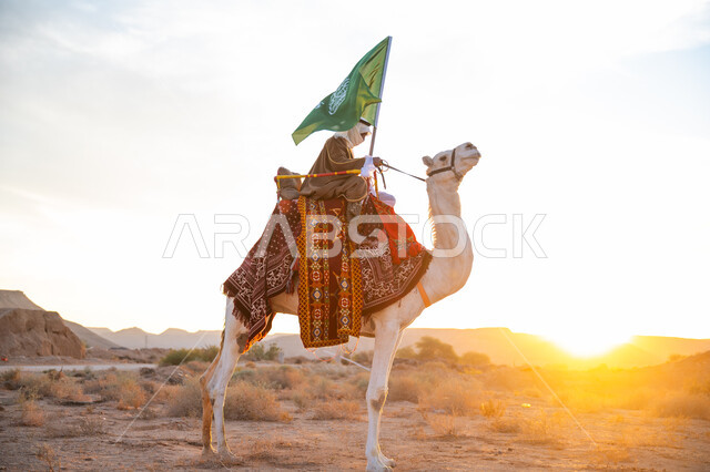 ارتداء الزي الشعبي بيوم تأسيس الدولة السعودية الأولى 1727 م, يوم بدينا 22 فبراير, الفخر والإنتماء للوطن، ارتباط الإبل بالتراث السعودي, رجل عربي خليجي سعودي يرتدي البشت والغترة يمتطي الجمل في الصحراء ويحمل علم المملكة بوقت الغروب, الاحتفال بعيد ومناسبة وطنية