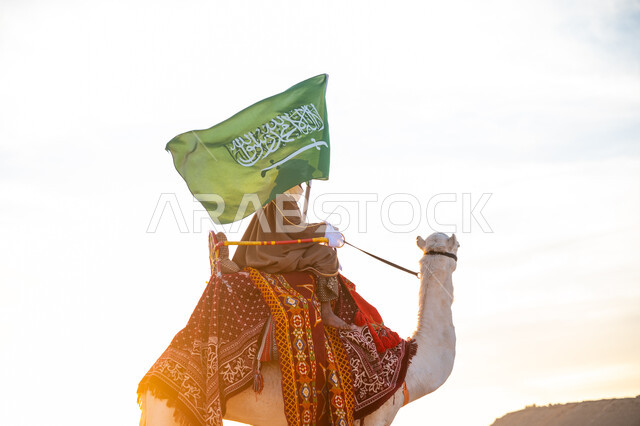 يوم بدينا 22 فبراير, الفخر والإنتماء للوطن، ارتباط الإبل بالتراث السعودي, رجل عربي خليجي سعودي يرتدي البشت والغترة يمتطي الجمل في الصحراء ويحمل علم المملكة, ارتداء الزي الشعبي بيوم تأسيس الدولة السعودية الأولى 1727 م, الاحتفال بعيد ومناسبة وطنية