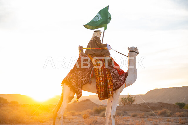 ارتداء الزي الشعبي بيوم تأسيس الدولة السعودية الأولى 1727 م, يوم بدينا 22 فبراير, الفخر والإنتماء للوطن، ارتباط الإبل بالتراث السعودي, رجل عربي خليجي سعودي يرتدي البشت والغترة يمتطي الجمل في الصحراء ويحمل علم المملكة بوقت الغروب, الاحتفال بعيد ومناسبة وطنية