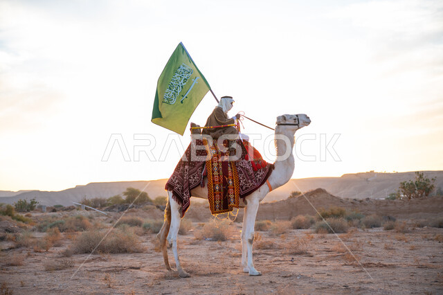 Anniversary of the founding of the first Saudi state 1727 AD, the day we started February 22, a Saudi Gulf Arab man wearing a bisht and a ghutra riding a camel in the desert and carrying the flag of the Kingdom, the banner of Islam and monotheism, wearing traditional folk costume on a national occasion, the concept of love of the homeland and belonging to it