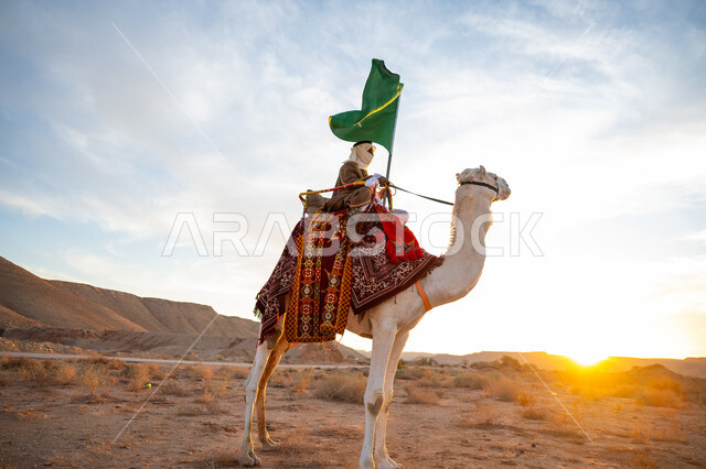 راية الإسلام والتوحيد, ارتداء الزي الشعبي التقليدي في مناسبة وطنية, مفهوم حب الوطن والانتماء اليه, يوم بدينا 22 فبراير, ذكرى تأسيس الدولة السعودية الأولى 1727م, رجل عربي خليجي سعودي يرتدي البشت والغترة يمتطي الجمل في الصحراء ويحمل علم المملكة بوقت غروب الشمس