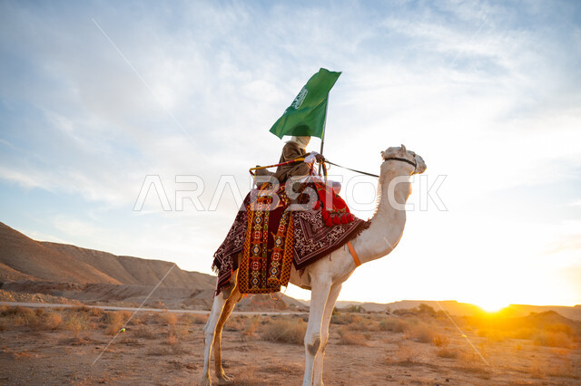 ذكرى تأسيس الدولة السعودية الأولى في 1727م, يوم بدينا 22 فبراير، ارتداء الزي الشعبي في المناسبة الوطنية, رجل عرب خليجي سعودي يرتدي البشت والغترة يمتطي الجمل في الصحراء ويحمل علم المملكة في وقت غروب الشمس, راية الإسلام والتوحيد, يوم العلم 11 مارس