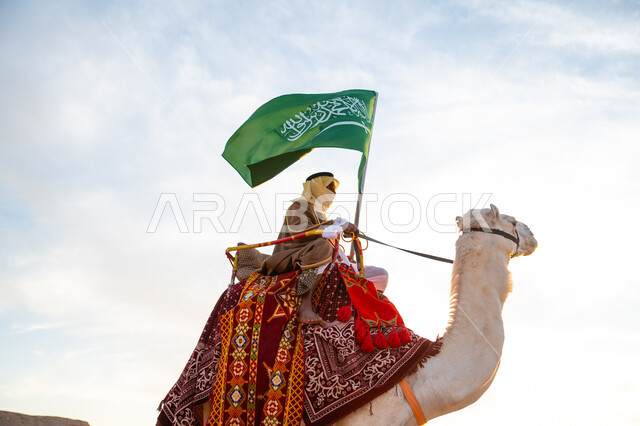 يوم العلم 11 مارس, ذكرى تأسيس الدولة السعودية الأولى في 1727م, يوم بدينا 22 فبراير، ارتداء الزي الشعبي في المناسبة الوطنية, صورة مقربة لرجل عرب خليجي سعودي يرتدي البشت والغترة يمتطي الجمل في الصحراء ويحمل علم المملكة, راية الإسلام والتوحيد