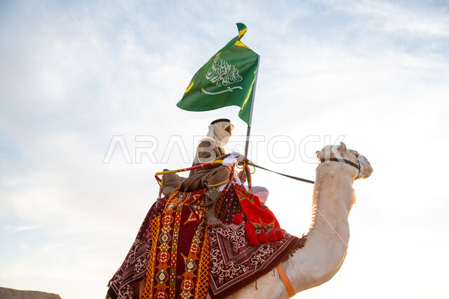 راية الإسلام والتوحيد, يوم العلم 11 مارس, ذكرى تأسيس الدولة السعودية الأولى في 1727م, يوم بدينا 22 فبراير، ارتداء الزي الشعبي في المناسبة الوطنية, صورة مقربة لرجل عرب خليجي سعودي يرتدي البشت والغترة يمتطي الجمل في الصحراء ويحمل علم المملكة