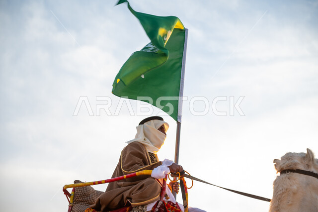 Flag Day March 11, the anniversary of the founding of the first Saudi state in 1727 AD, the day we started February 22, wearing the traditional costume on the national occasion, a close-up photo of a Saudi Gulf Arab man wearing a bisht and a ghutra riding a camel in the desert and carrying the flag of the Kingdom, the banner of Islam and monotheism