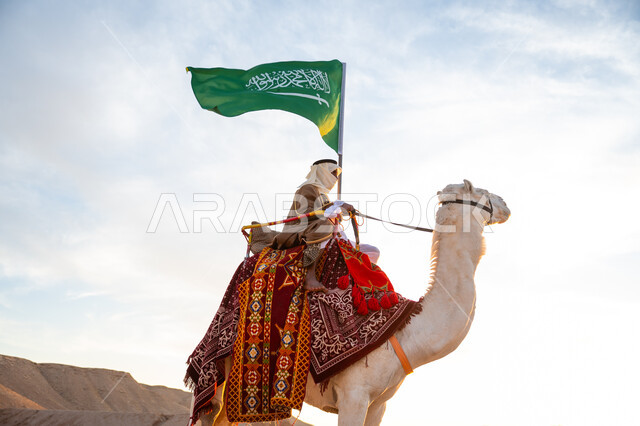 راية الإسلام والتوحيد, يوم العلم 11 مارس, ذكرى تأسيس الدولة السعودية الأولى في 1727م, يوم بدينا 22 فبراير، ارتداء الزي الشعبي في المناسبة الوطنية, صورة مقربة لرجل عرب خليجي سعودي يرتدي البشت والغترة يمتطي الجمل في الصحراء ويحمل علم المملكة