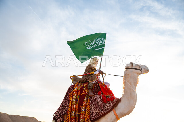 يوم بدينا 22 فبراير، ارتداء الزي الشعبي في المناسبة الوطنية, ذكرى تأسيس الدولة السعودية الأولى في 1727م, رجل عرب خليجي سعودي يرتدي البشت والغترة يمتطي الجمل في الصحراء ويحمل علم المملكة في وقت غروب الشمس, راية الإسلام والتوحيد, يوم العلم 11 مارس