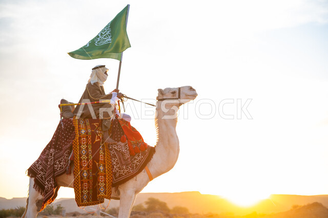 ذكرى تأسيس الدولة السعودية الأولى في 1727م, يوم بدينا 22 فبراير، ارتداء الزي الشعبي في المناسبة الوطنية, رجل عرب خليجي سعودي يرتدي البشت والغترة يمتطي الجمل في الصحراء ويحمل علم المملكة في وقت غروب الشمس, راية الإسلام والتوحيد, يوم العلم 11 مارس
