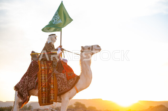 يوم العلم 11 مارس, ذكرى تأسيس الدولة السعودية الأولى في 1727م, يوم بدينا 22 فبراير، ارتداء الزي الشعبي في المناسبة الوطنية, رجل عرب خليجي سعودي يرتدي البشت والغترة يمتطي الجمل في الصحراء ويحمل علم المملكة في وقت غروب الشمس, راية الإسلام والتوحيد