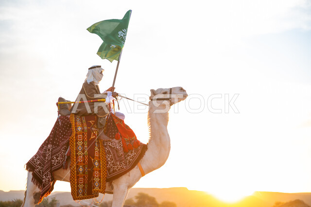راية الإسلام والتوحيد, يوم العلم 11 مارس, ذكرى تأسيس الدولة السعودية الأولى في 1727م, يوم بدينا 22 فبراير، ارتداء الزي الشعبي في المناسبة الوطنية, رجل عرب خليجي سعودي يرتدي البشت والغترة يمتطي الجمل في الصحراء ويحمل علم المملكة في وقت غروب الشمس