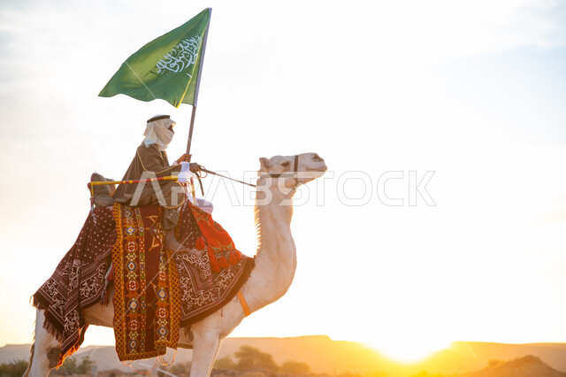يوم العلم 11 مارس, ذكرى تأسيس الدولة السعودية الأولى في 1727م, يوم بدينا 22 فبراير، ارتداء الزي الشعبي في المناسبة الوطنية, رجل عرب خليجي سعودي يرتدي البشت والغترة يمتطي الجمل في الصحراء ويحمل علم المملكة في وقت غروب الشمس, راية الإسلام والتوحيد