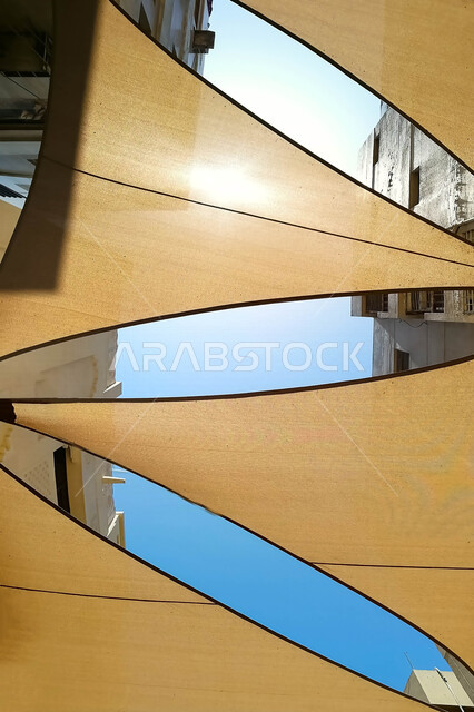 صورة لأشرعة على السقف بين المباني , مظلات قماشية تحمي من أشعة الشمس , تصاميم تشبه الشراع تعطي ظل للمكان .