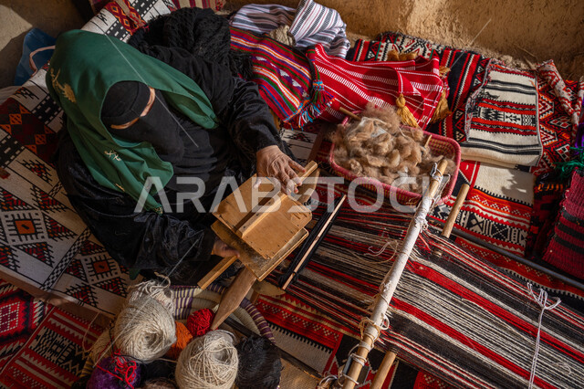 حرفة و مهنة يدوية تاريخيه, المحافظة على التراث السعودي، يوم بدينا 22 فبراير, ذكرى تأسيس الدولة السعودية الأولى 1727م, الاستعداد لنسج وغزل الخيوط, صورة من الاعلى لسيدة عربيه خليجية سعودية ترتدي العباءه السوداء و البرقع تقوم بتمشيط الصوف