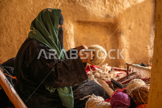 المحافظة على التراث الشعبي السعودي, يوم بدينا 22 فبراير, ذكرى تأسيس الدولة السعودية الأولى 1727م, حرفة يدوية تاريخيه, الشغف في ممارسة مهنة الغزل و النسج, صورة من الجانب لامرأة عربيه خليجية سعودية ترتدي البرقع و العباءه السوداء تقوم بلف خيوط الصوف