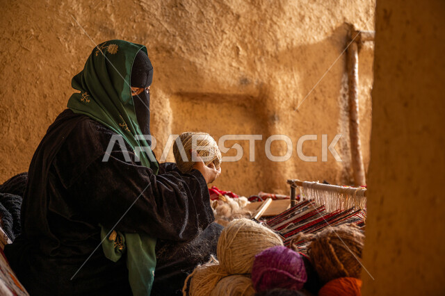 المحافظة على التراث الشعبي السعودي, يوم بدينا 22 فبراير, ذكرى تأسيس الدولة السعودية الأولى 1727م, حرفة يدوية تاريخيه, الشغف في ممارسة مهنة الغزل و النسج, صورة من الجانب لامرأة عربيه خليجية سعودية ترتدي البرقع و العباءه السوداء تقوم بلف خيوط الصوف